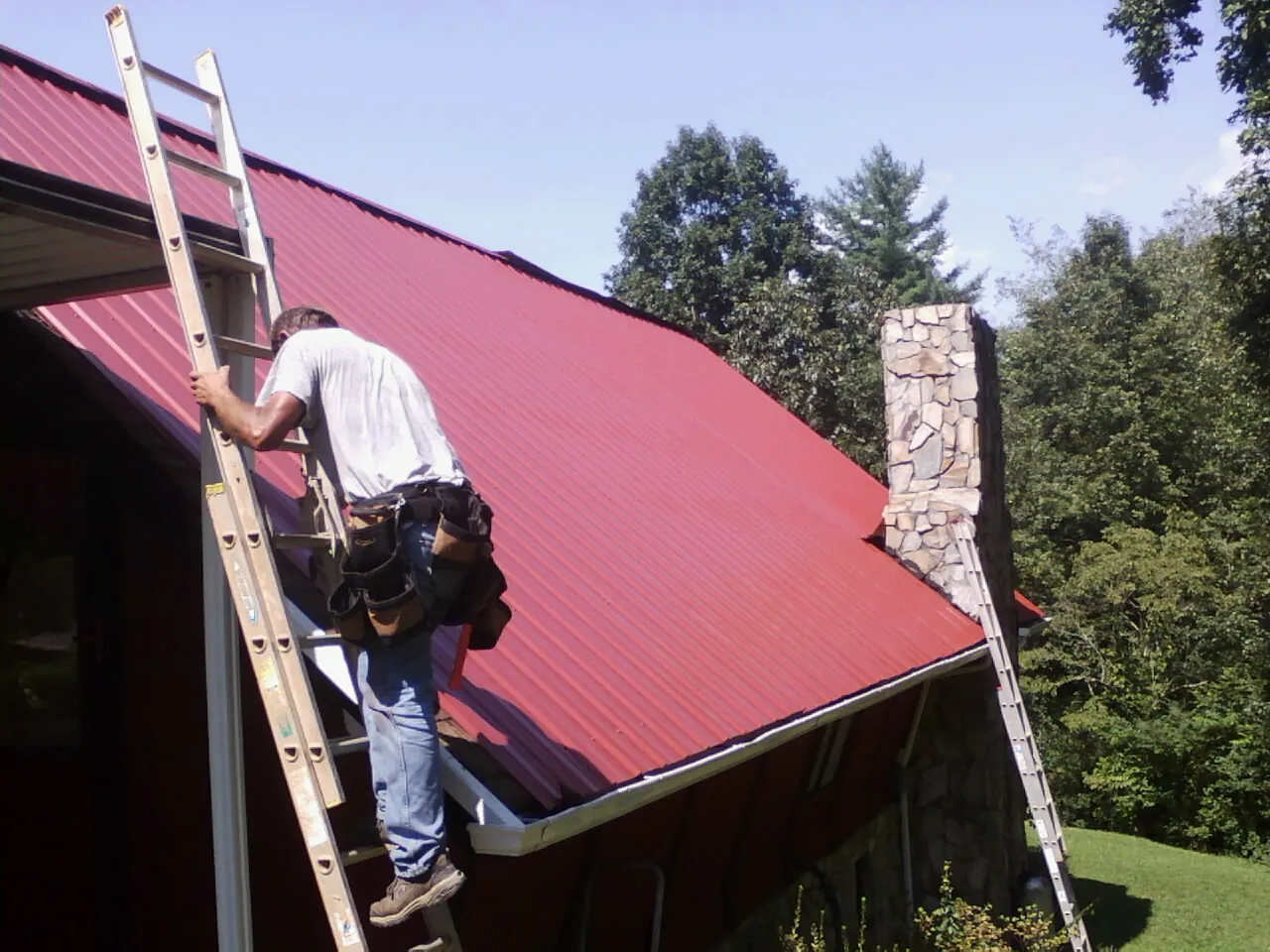 Roofer on a ladder beside a red metal roof and stone chimney — Soffit & Fascia Repair by Roofing Brothers in Slatersville